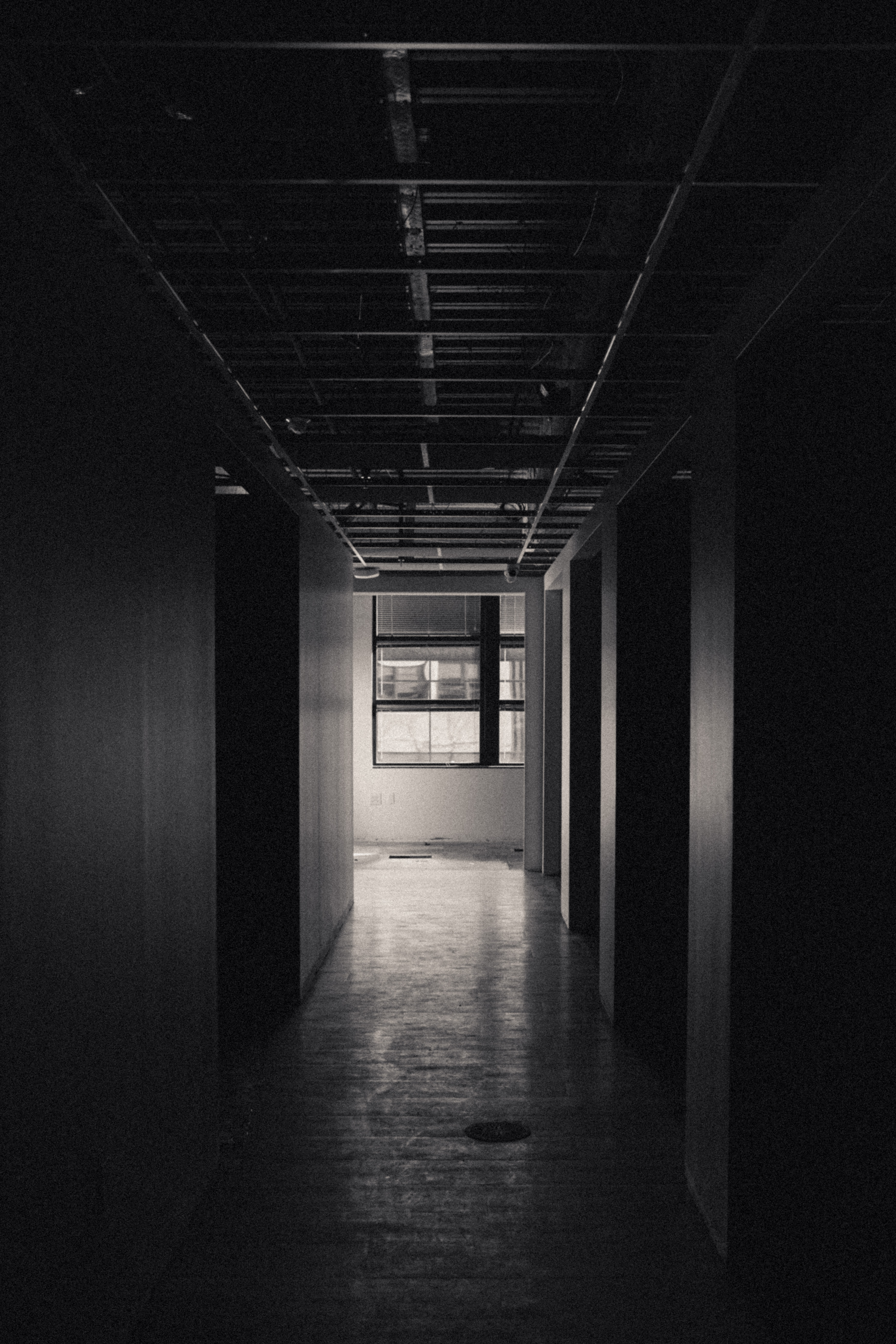 “Black and white photo looking down an office hallway toward a window. There are dark office spaces along the sides of the hallway. Doors, lights, and ceiling tiles have not been installed yet.”